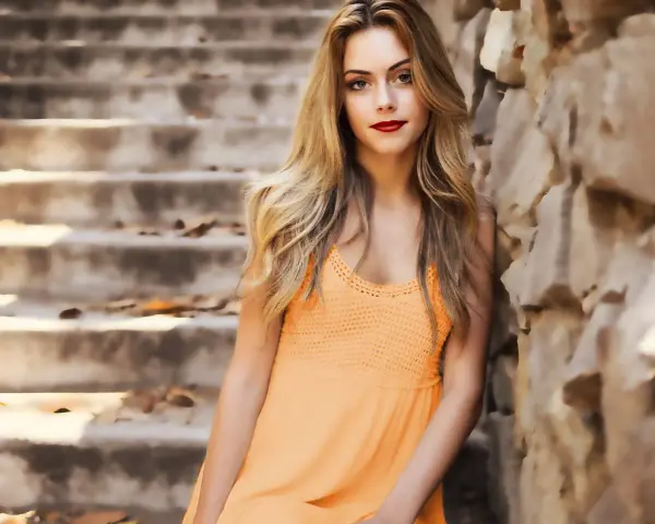 Jeune femme blonde, robe orange, appuyée contre un mur de pierres, escalier en arrière-plan.