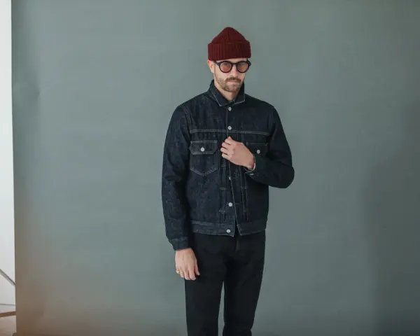 Homme portant une veste en jean sombre et un pantalon noir, bonnet bordeaux et lunettes rondes.