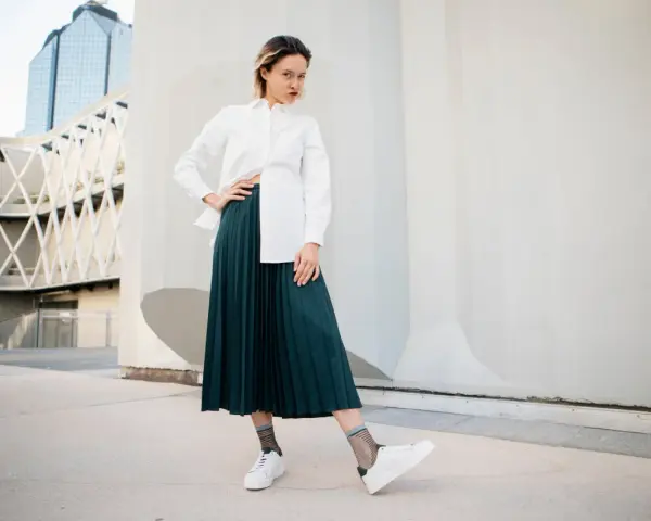 Une femme pose avec confiance, portant une chemise blanche et une élégante jupe plissée vert foncé.