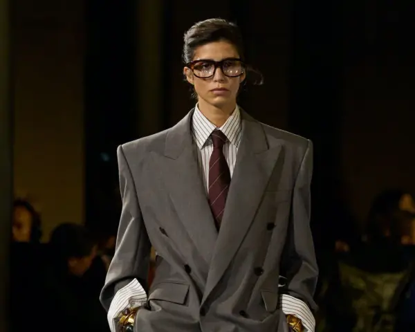 Mannequin au style collegienne, portant un costume gris oversize, chemise à rayures et cravate bordeaux. Lunettes imposantes.