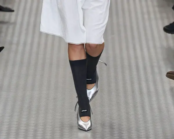 Une mannequin défile avec une tenue blanche et des chaussettes hautes grises, rehaussée par des chaussures argentées pointues.