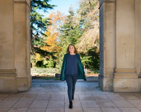 Une femme souriante marche sous une arche, portant un manteau vert et un haut bleu marine. L'automne colore les arbres en arrière-plan.