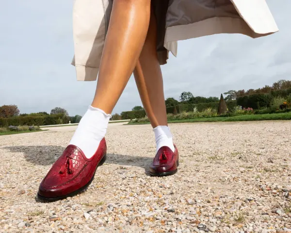 Pieds chaussés de mocassins rouges vernis, ornés de pompons, marchant sur un chemin de gravier. Le rouge vif de ces chaussures suggère audace et passion.