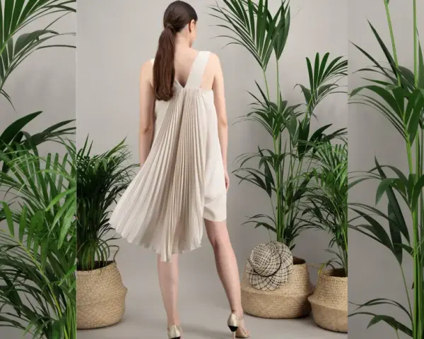 Une femme porte une robe plissée beige, parfaite pour une tenue mariage septembre. Elle est entourée de plantes vertes luxuriantes dans des paniers en osier.