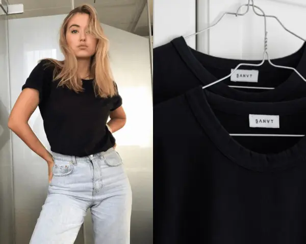 Une jeune femme porte une tenue avec t-shirt noir et jean clair. Deux t-shirts noirs identiques sont accrochés sur des cintres.
