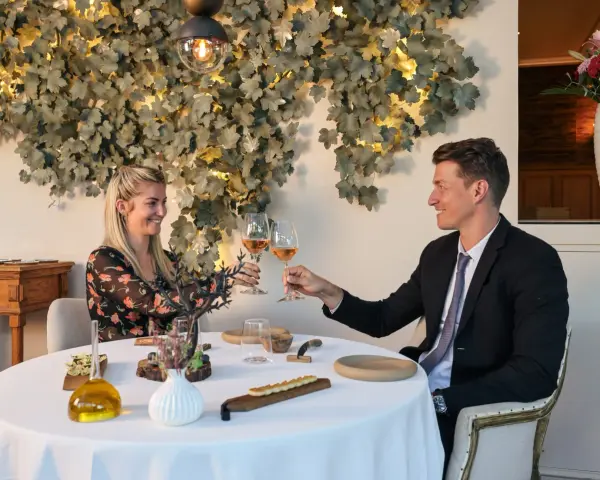 Un couple trinque avec du vin rosé dans un restaurant gastronomique, décoré de feuilles stylisées et d'une lampe design.