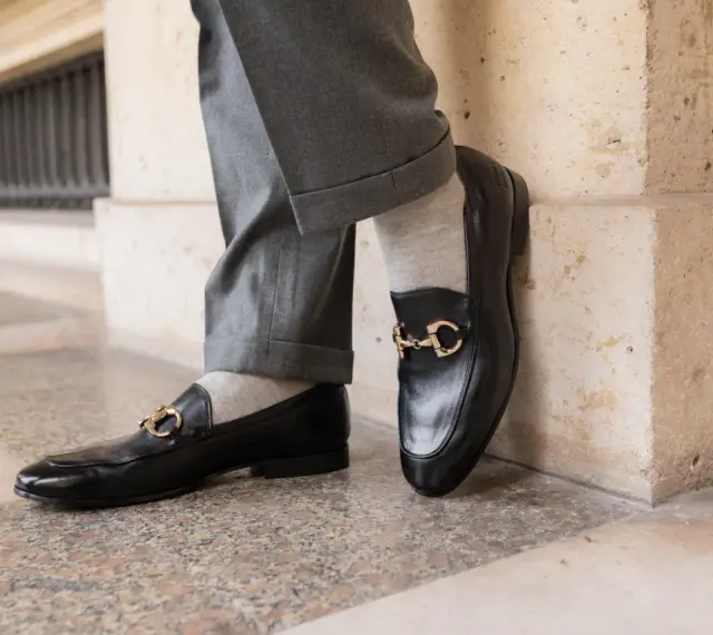 Un homme porte un pantalon gris retroussé et une chaussette mocassin noire avec une boucle dorée.