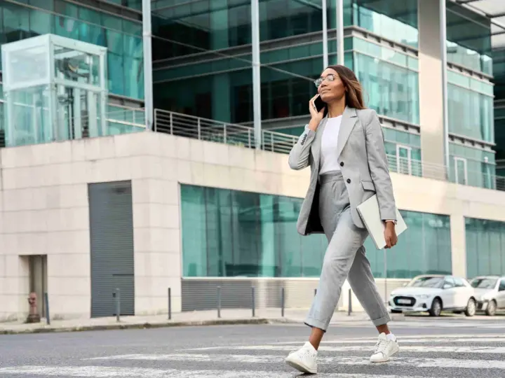 Femme en tenue d'entretien d'embauche d'été, tailleur gris, traversant la rue avec un ordinateur portable, parlant au téléphone.