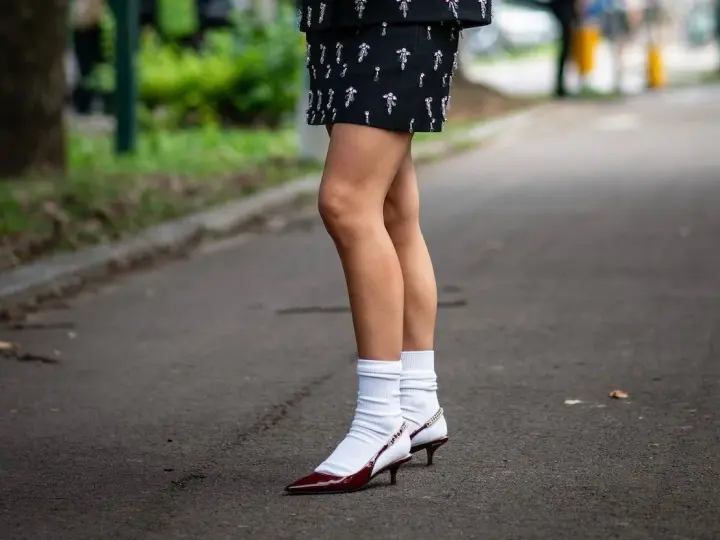 Jambes portant une jupe ornée et des escarpins bordeaux avec une chaussette par dessus chaussure blanche.