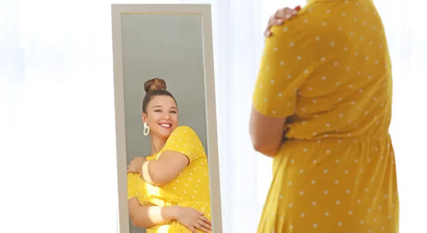 Une femme se regarde dans un miroir, souriante, portant une robe jaune à pois. Elle semble heureuse, malgré la robe trop courte.