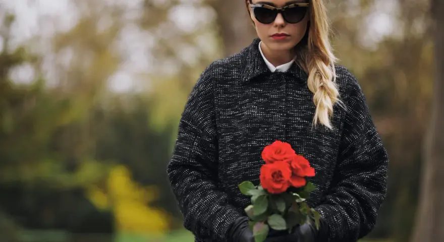 Une jeune femme, portant une tenue d'enterrement hiver, tient un bouquet de roses rouges. Ses lunettes de soleil cachent ses yeux, mais son expression est empreinte de tristesse.