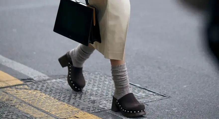 Une femme marche dans la rue, portant un sac noir et un élégant sabot femme marron clouté avec des chaussettes grises.
