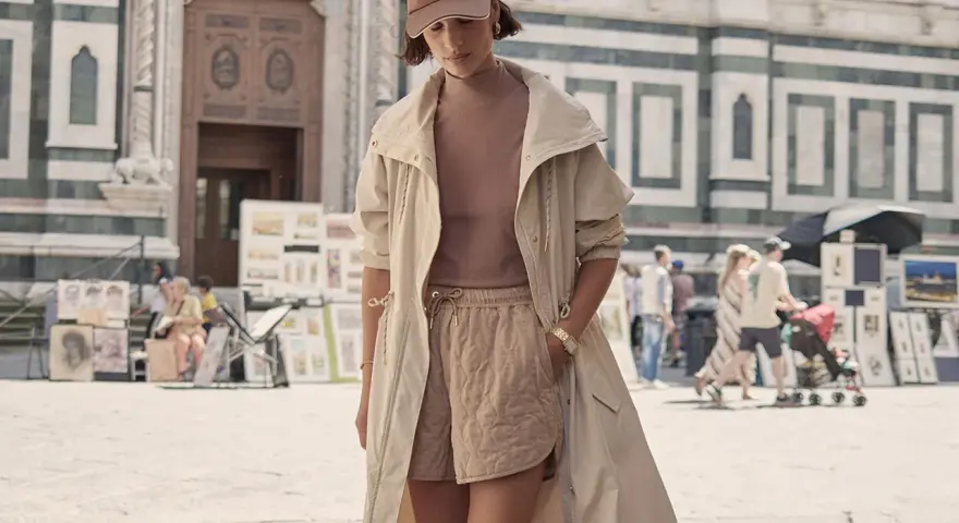 Une femme porte un ensemble athleisure chic : short matelassé, haut col roulé, veste longue et casquette. Elle se promène devant une cathédrale.
