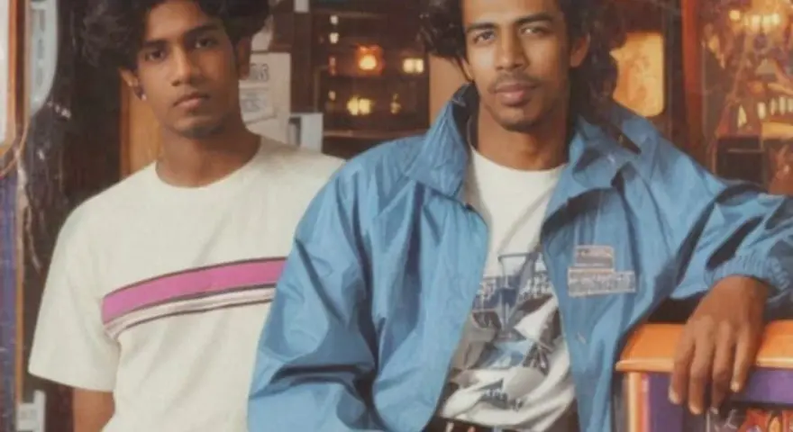 Deux jeunes hommes posent, arborant un style vestimentaire année 80 : t-shirt à rayures roses et veste bleue.