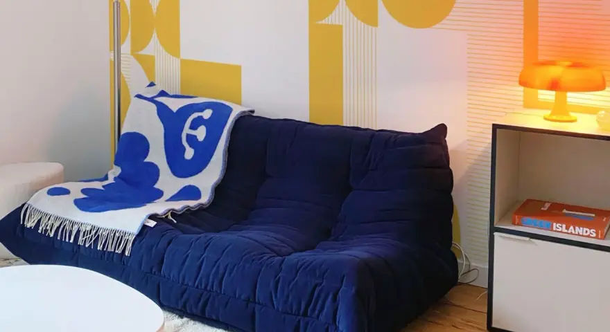 Canapé bleu profond avec plaid blanc et bleu, sur fond de papier peint jaune géométrique en **color blocking**. Lampe orange sur une étagère.