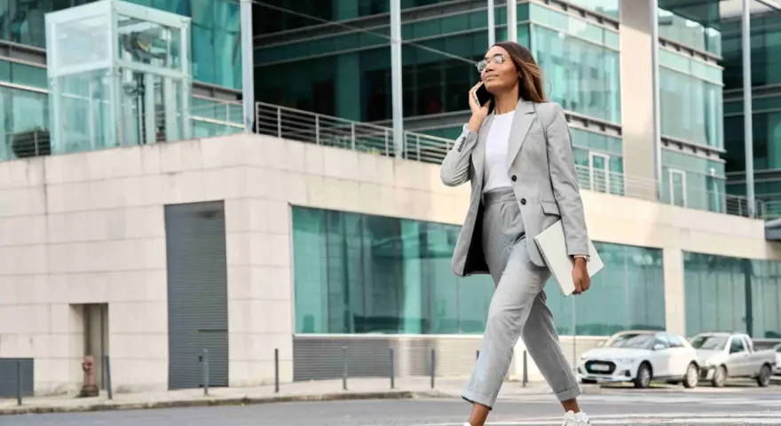 Femme en tenue d'entretien d'embauche d'été, tailleur gris, traversant la rue avec un ordinateur portable, parlant au téléphone.