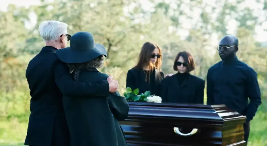 Un couple en tenue d'enterrement, le bras autour de l'épaule, se tient près d'un cercueil. D'autres personnes en deuil sont derrière.