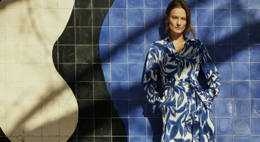 Une femme pose devant un mur de carreaux bleus et blancs. Sa robe à motifs bleus et blancs s'harmonise parfaitement avec le décor. Quelle couleur va bien avec le bleu vêtement ? Le blanc, évidemment !