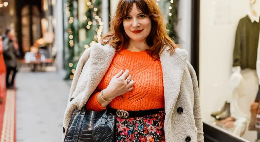Une femme arbore une tenue avec pull orange vif, jupe fleurie et manteau douillet, prête pour une sortie festive.