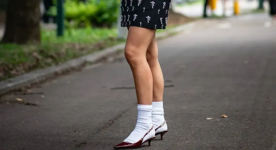 Jambes portant une jupe ornée et des escarpins bordeaux avec une chaussette par dessus chaussure blanche.