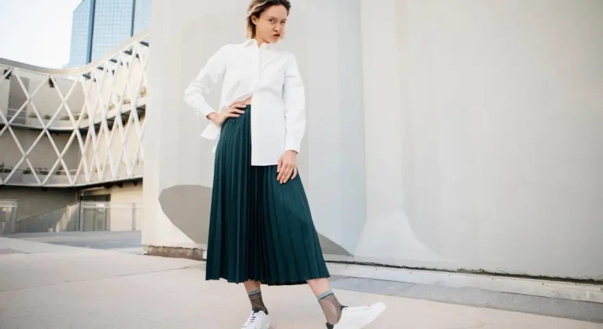 Une femme pose avec confiance, portant une chemise blanche et une élégante jupe plissée vert foncé.