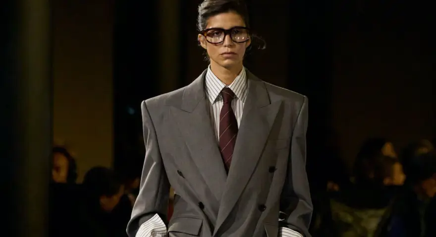 Mannequin au style collegienne, portant un costume gris oversize, chemise à rayures et cravate bordeaux. Lunettes imposantes.