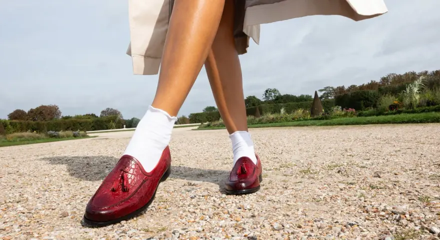 Pieds chaussés de mocassins rouges vernis, ornés de pompons, marchant sur un chemin de gravier. Le rouge vif de ces chaussures suggère audace et passion.