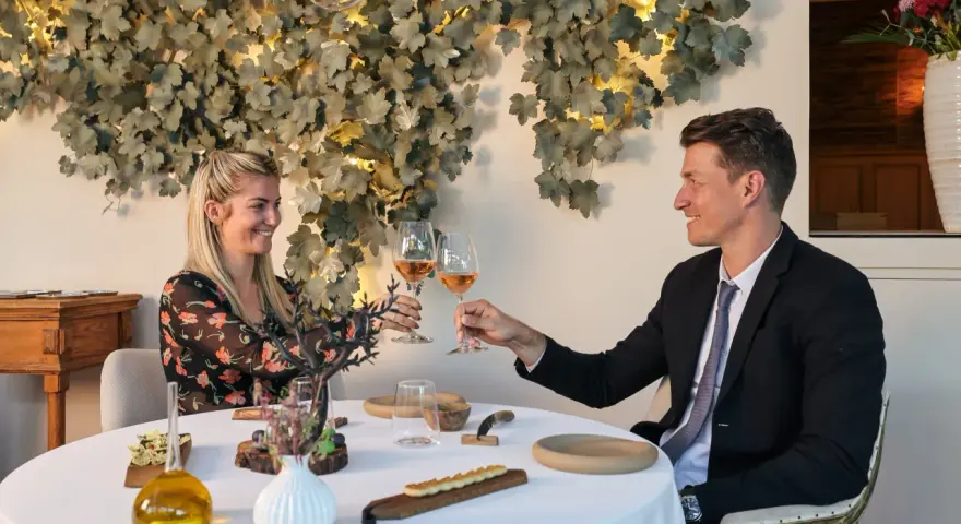 Un couple trinque avec du vin rosé dans un restaurant gastronomique, décoré de feuilles stylisées et d'une lampe design.