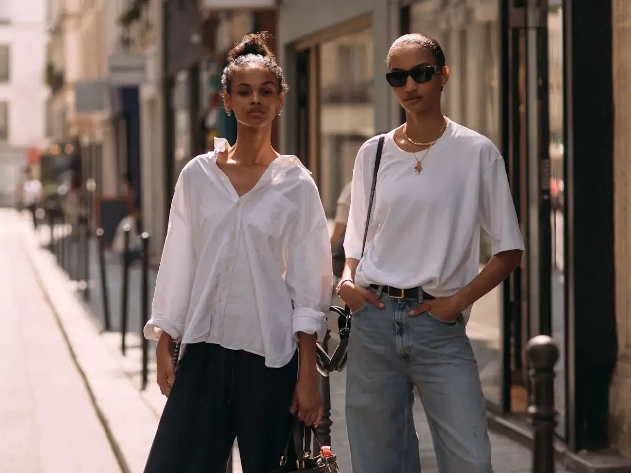 Deux femmes affichent un style vestimentaire streetwear décontracté dans une rue parisienne. L'une porte une chemise blanche ample et un pantalon noir, l'autre un t-shirt blanc et un jean.