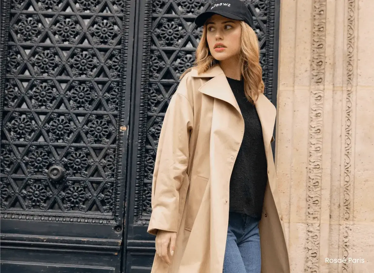Une look femme 40 ans moderne d&eacute;contract&eacute;e, portant un trench beige, un pull noir et un jean, devant une porte ornement&eacute;e.