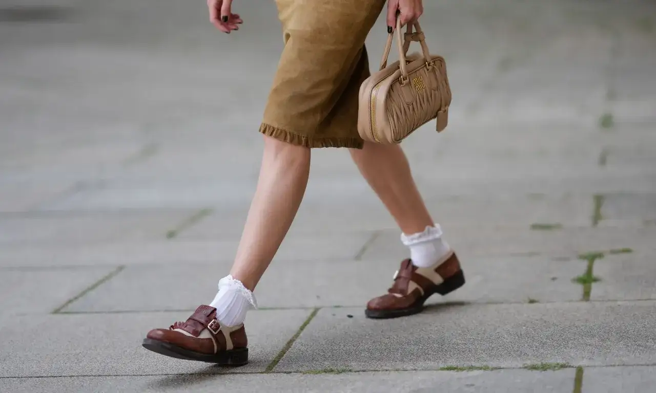 Une femme porte une jupe en daim marron, des chaussures marron et beige avec une chaussette par dessus chaussure blanche &agrave; dentelle. Elle tient un sac Miu Miu beige.