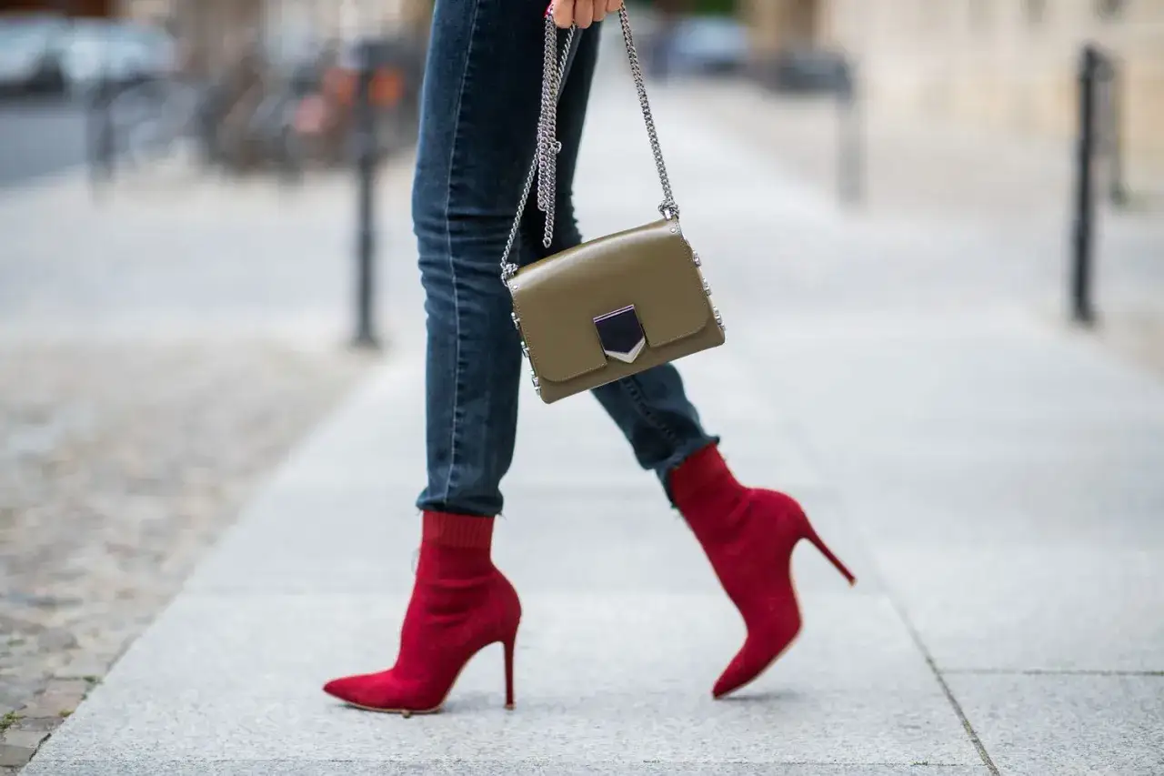 Une femme porte un jean slim et des bottines rouges à talons aiguilles, tenant un sac à main kaki.
