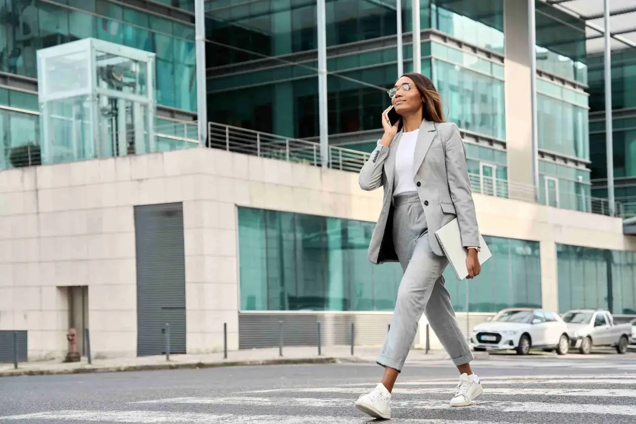Une femme en tenue d'entretien d'embauche d'été, tailleur gris, chemise blanche et baskets, traverse la rue en parlant au téléphone, un ordinateur portable à la main.
