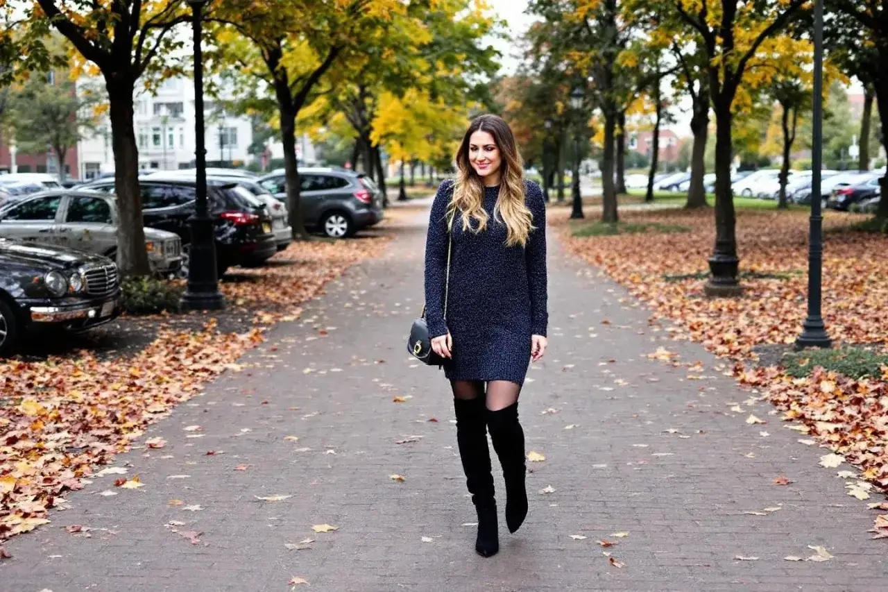 Une femme porte une robe noire et des bottes hautes, id&eacute;ale pour l'hiver. Le chemin est jonch&eacute; de feuilles d'automne.