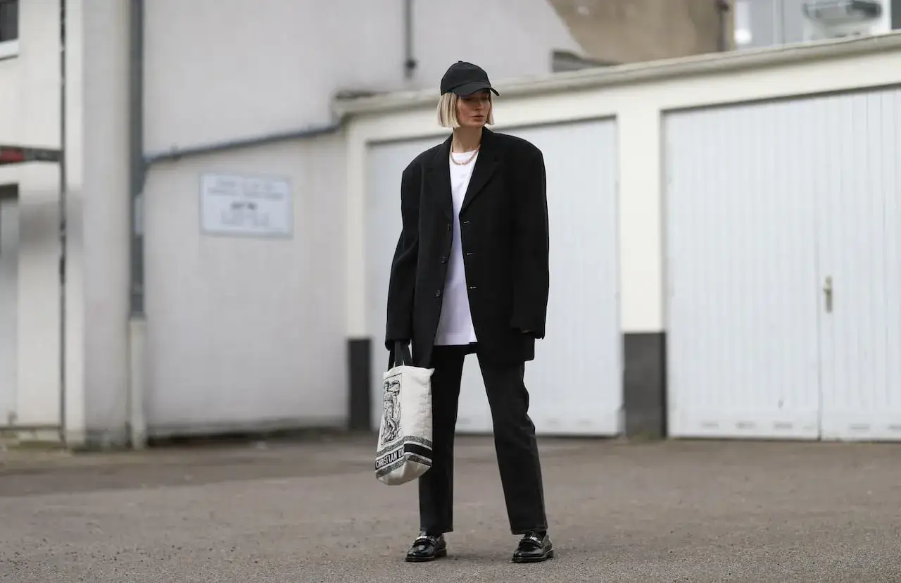 Une femme porte une chemise blanche, un pantalon noir, un blazer oversize et une casquette. Elle tient un sac en toile.