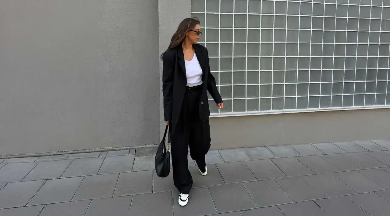 Une femme porte un &eacute;l&eacute;gant outfit pull sans manche noir, un blazer oversize et un sac &agrave; main. Elle marche sur un trottoir devant un mur gris.