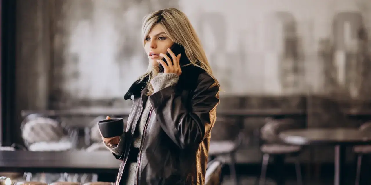Femme blonde en veste en cuir, tenant une tasse et parlant au téléphone dans un café.