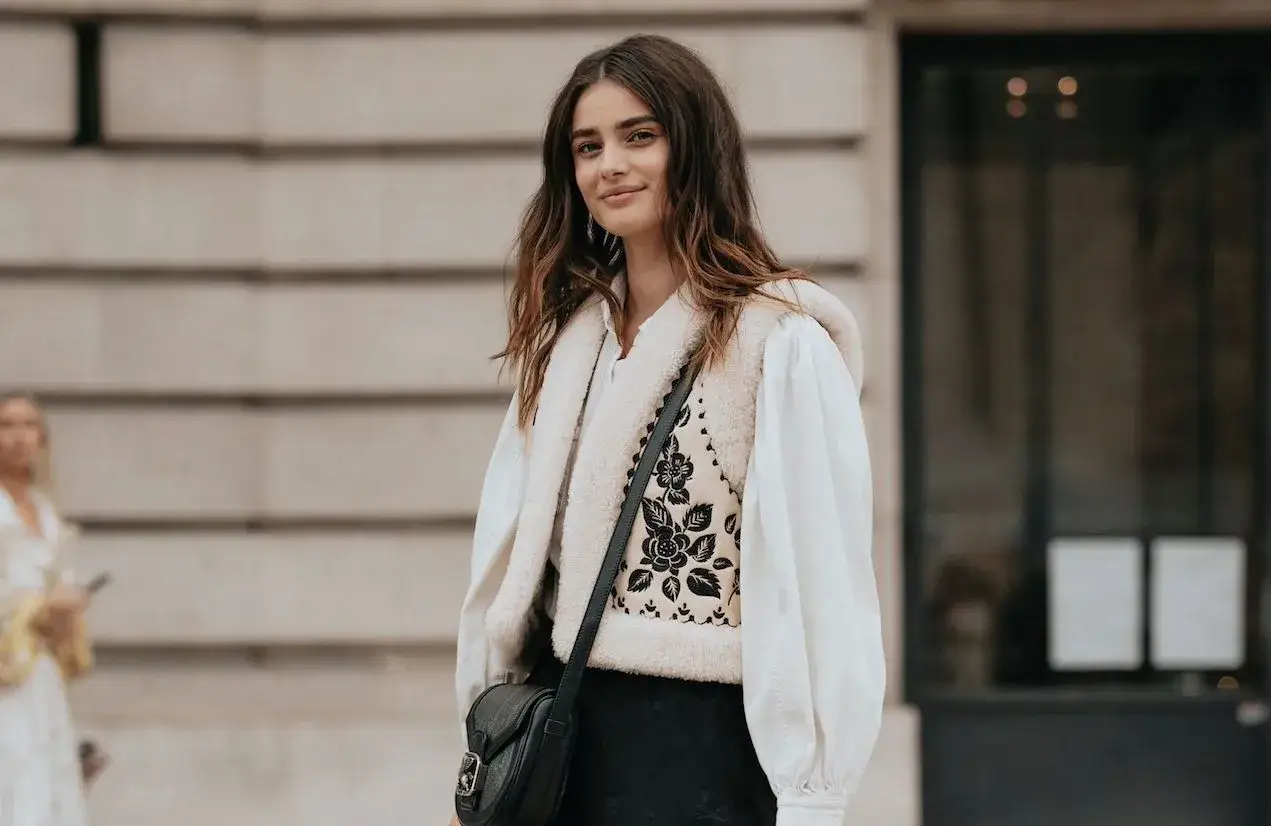 Une jeune femme au style bobo, portant un gilet en fausse fourrure sur une chemise blanche et un pantalon noir.