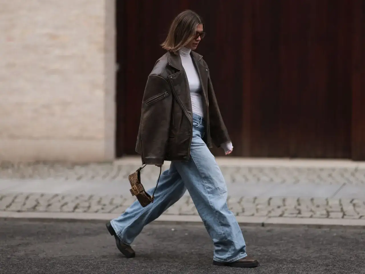 Une femme porte une veste en cuir marron, un col roulé blanc et un jean large, un sac à main monogrammé à la main.