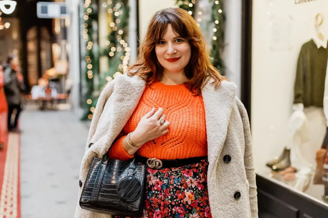 Une femme arbore une tenue avec pull orange vif, jupe fleurie et manteau douillet, pr&ecirc;te pour une sortie festive.