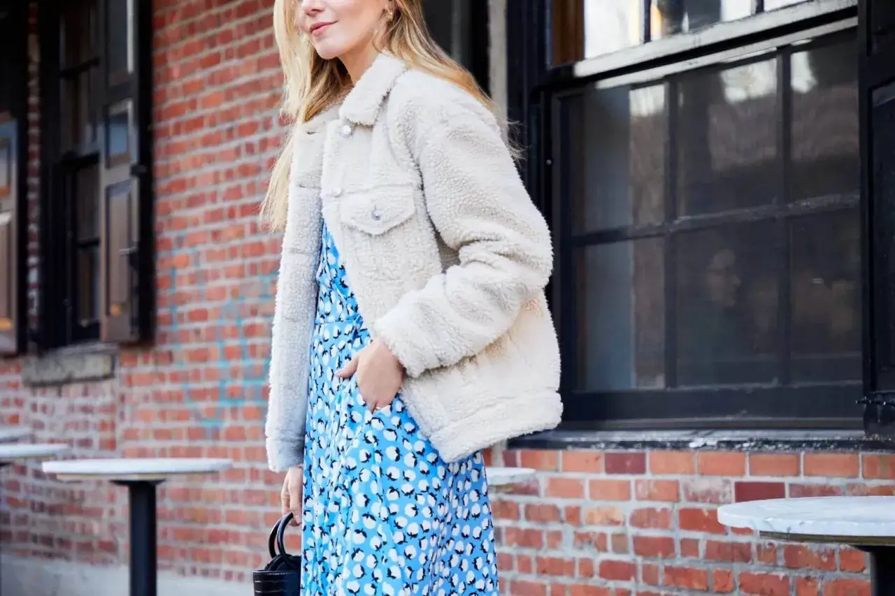 Une femme porte une robe courte bleue &agrave; motifs blancs et une veste en sherpa cr&egrave;me. Une id&eacute;e parfaite pour savoir quelle veste porter avec une robe courte.