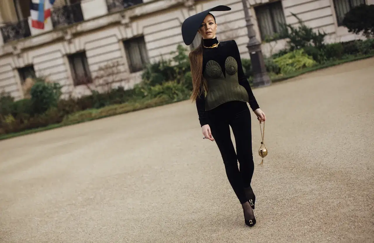 Une femme porte une tenue avec legging noir, un corset vert olive et un chapeau noir audacieux, tenant un sac doré.