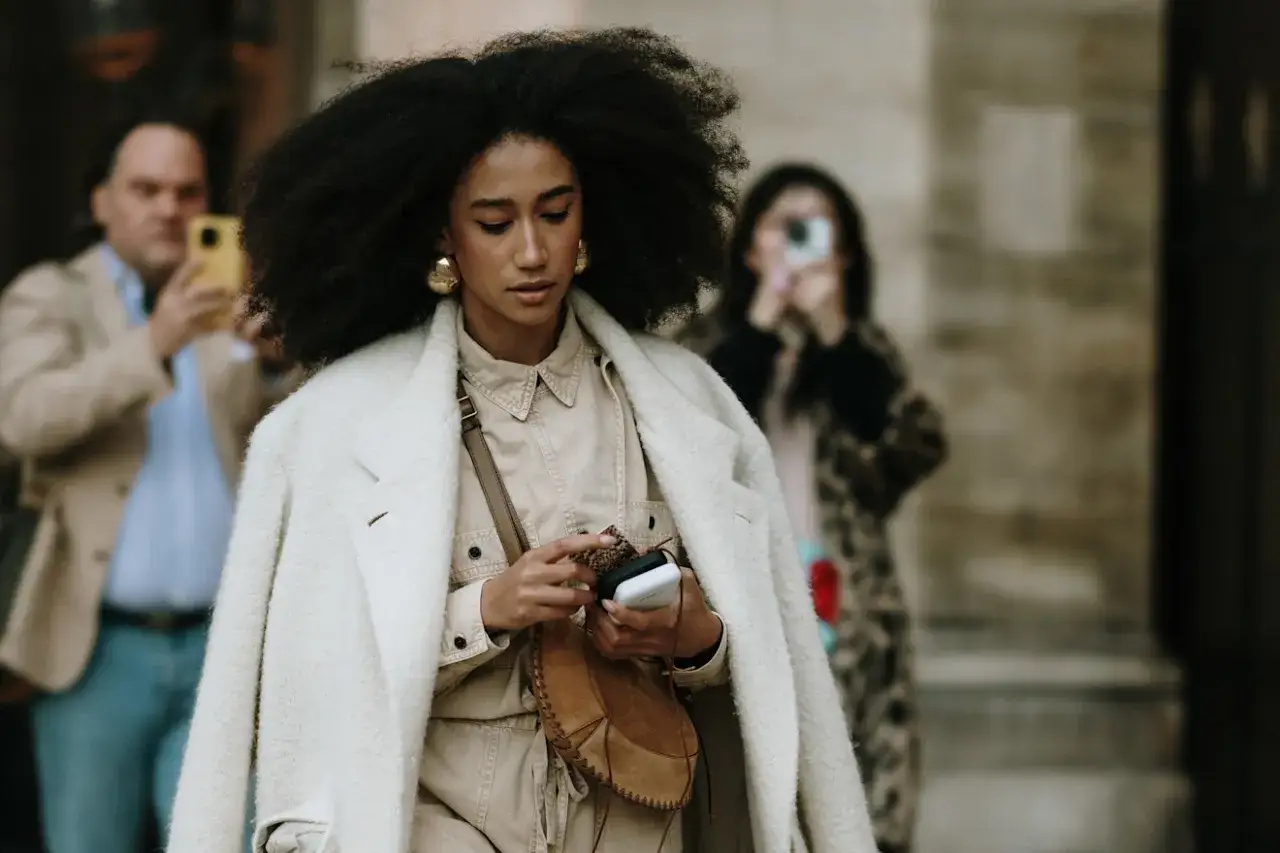 Une femme au style bohème chic, avec une chevelure afro volumineuse, porte un manteau crème et une tenue beige. Elle manipule son téléphone, un sac en bandoulière en daim à ses côtés.