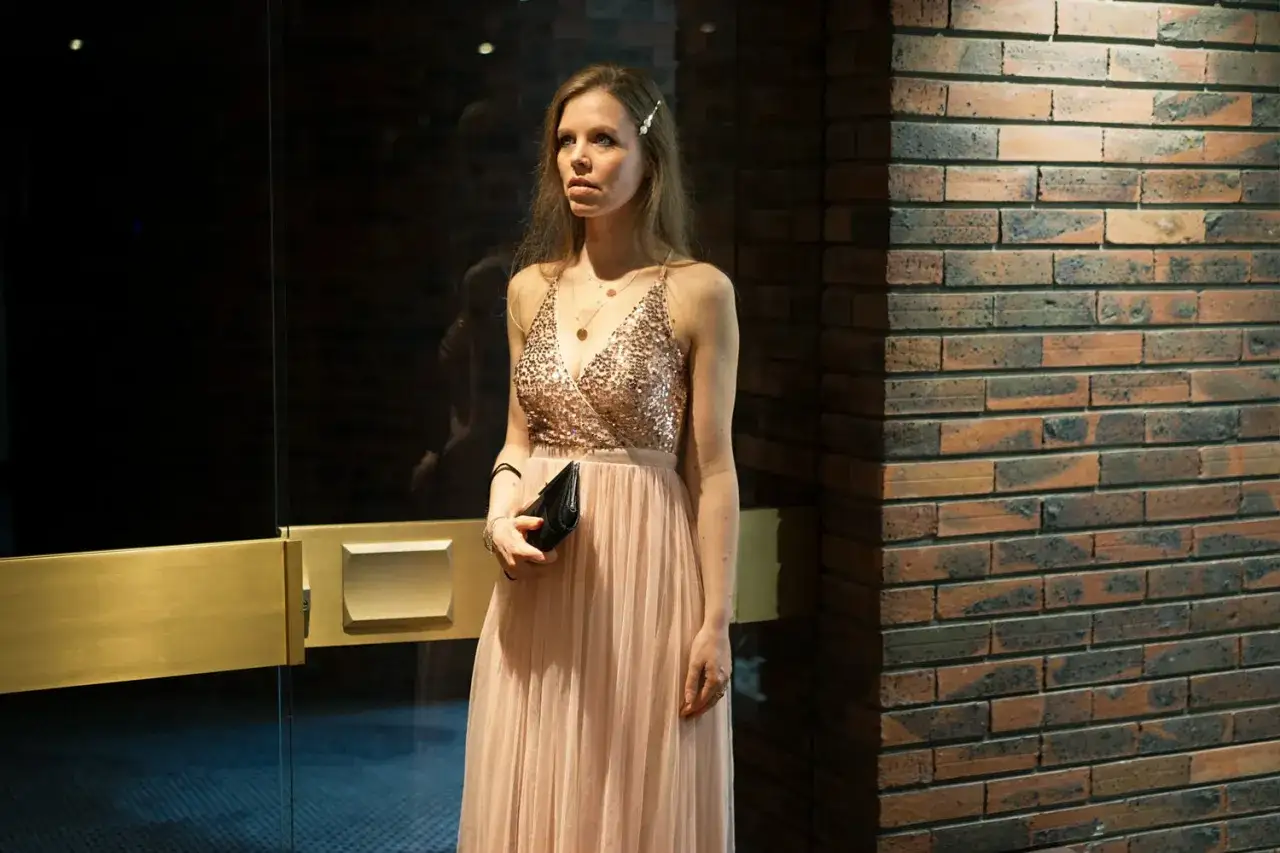 Une femme &eacute;l&eacute;gante, dans sa tenue d'op&eacute;ra, attend devant une porte vitr&eacute;e. Sa robe longue et l&eacute;g&egrave;re, orn&eacute;e de sequins, brille sous la lumi&egrave;re.
