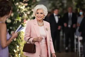 Élégante femme de 75 ans, radieuse dans sa tenue de mariage rose poudré, collier de perles et broche scintillante.