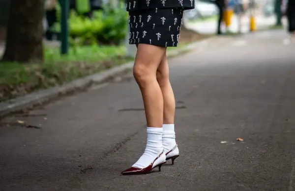 Jambes portant une jupe ornée et des escarpins bordeaux avec une chaussette par dessus chaussure blanche.