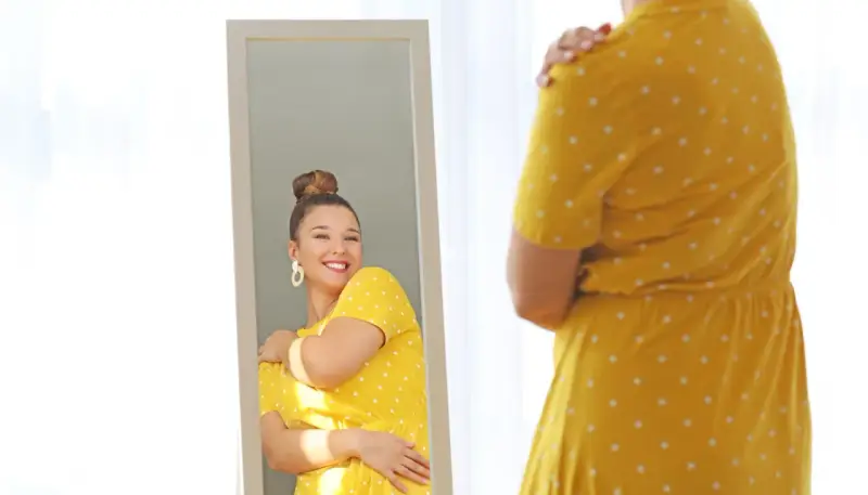 Une femme se regarde dans un miroir, souriante, portant une robe jaune à pois. Elle semble heureuse, malgré la robe trop courte.