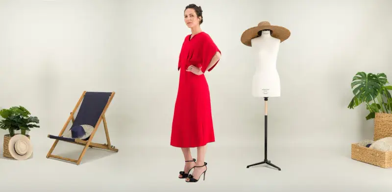 Une femme pose dans une robe rouge éclatante. À ses côtés, une chaise longue, un mannequin de couture et des plantes. Une robe casual, c'est quoi ?