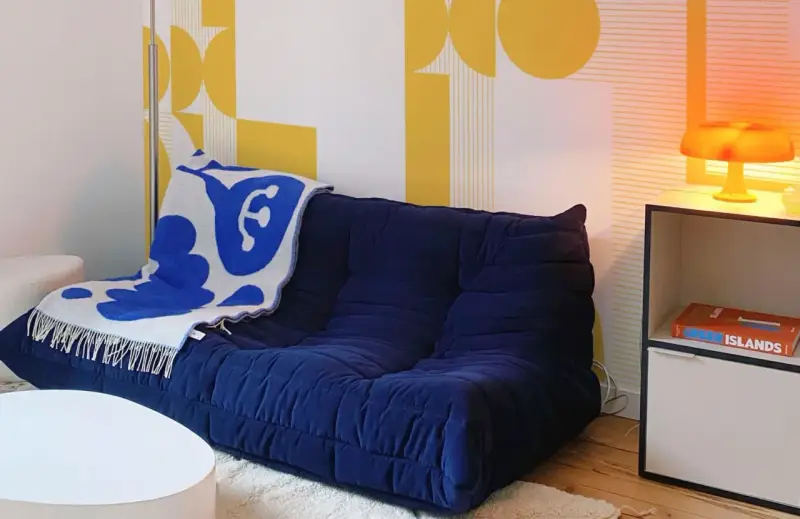 Canapé bleu profond avec plaid blanc et bleu, sur fond de papier peint jaune géométrique en **color blocking**. Lampe orange sur une étagère.