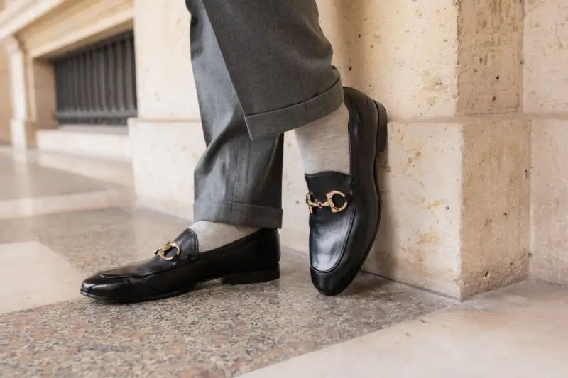 Un homme porte un pantalon gris retroussé et une chaussette mocassin noire avec une boucle dorée.